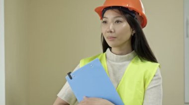 Asian woman builder in protective clothing. A young woman is holding a folder with documents and smiling.