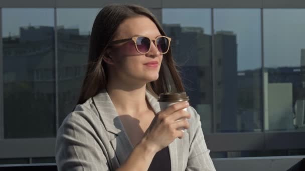 Smiling young woman in sunglasses drinks coffee from a disposable cup. — Stockvideo