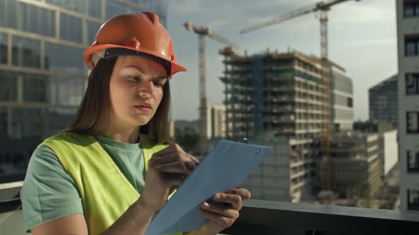 Młoda budowniczka w kamizelce sygnalizacyjnej i kasku coś pisze. Na tle budowanego budynku. — Wideo stockowe