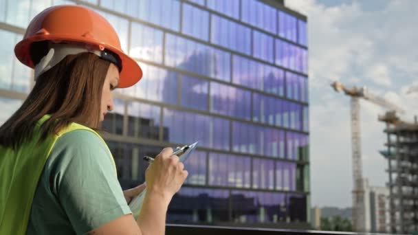 Young female builder in a signal vest and helmet writes something. Against the backdrop of a building under construction. — Αρχείο Βίντεο