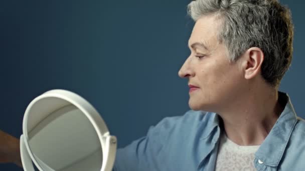 An elderly woman who cares about her appearance tints her eyes while sitting in front of a mirror at home. — Vídeo de Stock