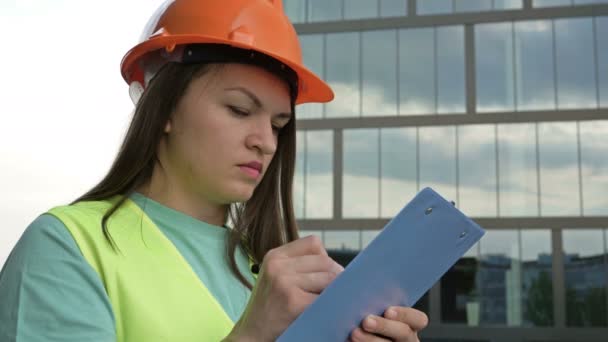 Young female builder in a signal vest and helmet writes something. Against the backdrop of a building under construction. — Αρχείο Βίντεο