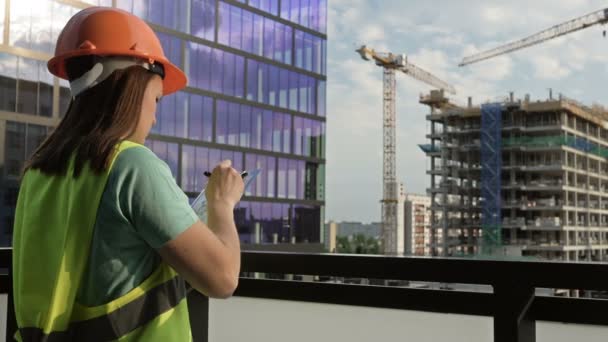 穿着信号弹背心和头盔的年轻女建筑工人写了一些东西。在一幢正在建造的大楼的背景下. — 图库视频影像