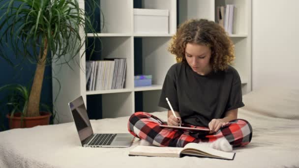 Мила дівчина-підліток робить домашнє завдання, сидячи на ліжку вдома. Молода красива дівчина думає і пише щось в блокноті, навчається онлайн, дистанційне навчання, самоосвіта . — стокове відео