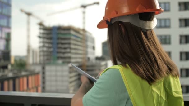 Vrouwelijke bouwer in een signaalvest en helm schrijft iets. Tegen de achtergrond van een gebouw in aanbouw. — Stockvideo