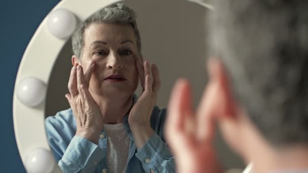 Eine ältere, ergraute Frau untersucht sorgfältig ihr Spiegelbild im Spiegel und berührt leicht die Falten auf der Haut ihres Gesichts.. — Stockvideo