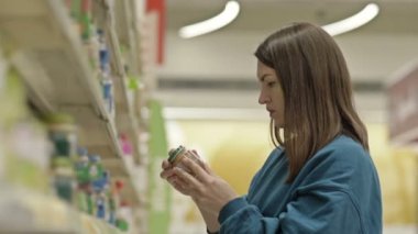 Genç bayan müşteri, markette bebek mamasıyla birlikte rafların yanındaki kavanozdaki etiketi dikkatle inceliyor. 4K.