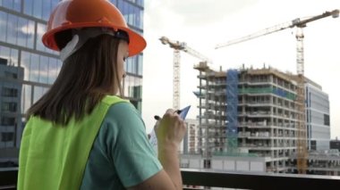 Young female builder in a signal vest and helmet writes something. Against the backdrop of a building under construction. HD.