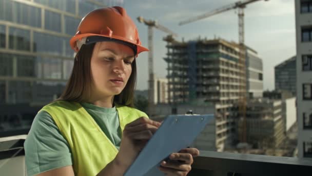 穿着信号弹背心和头盔的年轻女建筑工人写了一些东西 在一座正在建造的大楼的背景下 — 图库视频影像