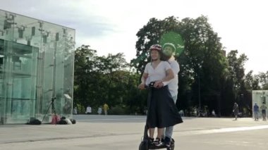 Genç adam şehir meydanında kız arkadaşına elektrikli scooterla biniyor. İkisi de koruyucu kask takıyor. 4K.