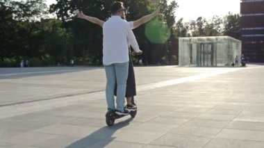 Genç adam şehir meydanında elektrikli scooterla kız arkadaşına biniyor. Mutlu kız gülüyor ve kollarını yana açıyor. 4K.