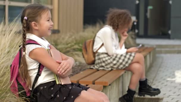 異なる年齢の2人の女子学生が学校の庭のベンチに座っています 少女は本を胸に押し当て 長老はスマートフォンを手にしている — ストック動画