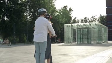 Genç adam şehir meydanında kız arkadaşına elektrikli scooterla biniyor. İkisi de koruyucu kask takıyor. HD.