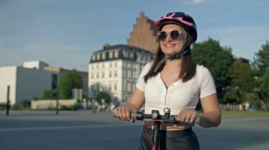Güneş gözlüklü ve kasklı genç bir kadın güzel bir şehir meydanının ortasında elektrikli scooterın üzerinde duruyor. HD.