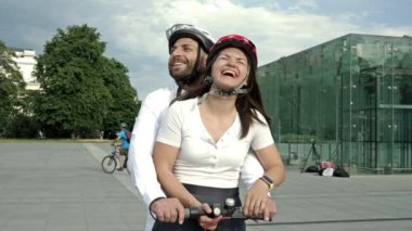 Genç adam şehir meydanında elektrikli scooterla kız arkadaşına biniyor. İkisi de mutlu bir şekilde gülümser HD.