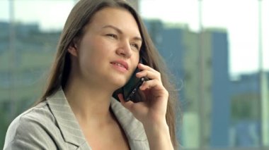 Beautiful young woman talking on the phone while standing against the backdrop of an office window. HD.