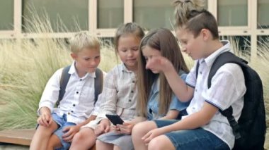 Group of elementary school students sit on a bench in the schoolyard. Children with interest look at the screen of a smartphone in the hands of a girl. 4K.