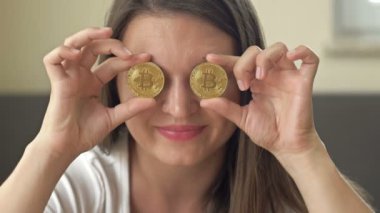 Cheerful woman playfully covers her eyes with gold coins - bitcoins. 4K.