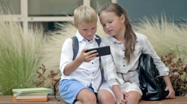 Two friends, a boy and a girl aged 8-9, are sitting on a bench in the schoolyard. Children look at the smartphone screen with interest. 4K.