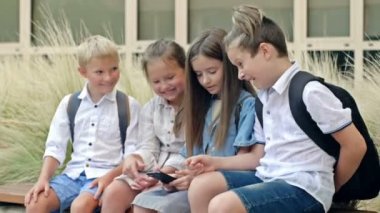 Group of elementary school students sit on a bench in the schoolyard. Children with interest look at the screen of a smartphone in the hands of a girl. HD.