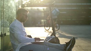 Concept of remote work. A middle-aged man in headphones sits on the sidewalk in the city and works with a laptop and smartphone while drinking coffee. Electric scooter nearby. HD.