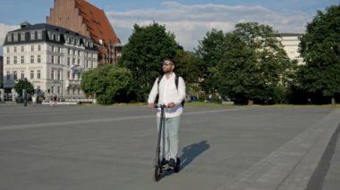 Bearded man with a backpack and in sunglasses rides an electric scooter through the city square. The concept of convenient and environmentally friendly individual urban transport. HD.