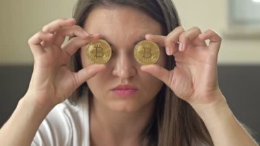 Cheerful woman playfully covers her eyes with gold coins - bitcoins. 4K.