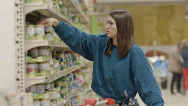 若い女性の顧客は慎重にスーパーマーケットでベビーフード付き棚の近くの瓶のラベルを調べます — ストック動画