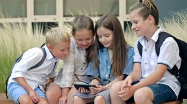 Group of elementary school students sit on a bench in the schoolyard. Children with interest look at the screen of a smartphone in the hands of a girl. 4K.