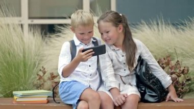 Two friends, a boy and a girl aged 8-9, are sitting on a bench in the schoolyard. Children look at the smartphone screen with interest. 4K.