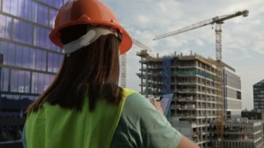 Female builder in a signal vest and helmet writes something. Against the backdrop of a building under construction. Hd.