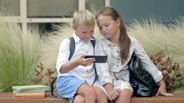 Two friends, a boy and a girl aged 8-9, are sitting on a bench in the schoolyard. Children look at the smartphone screen with interest. 4K.