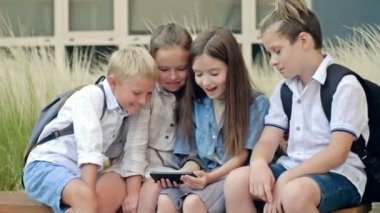 Group of elementary school students sit on a bench in the schoolyard. Children with interest look at the screen of a smartphone in the hands of a girl. HD.