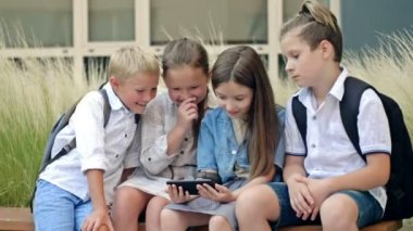 Group of elementary school students sit on a bench in the schoolyard. Children with interest look at the screen of a smartphone in the hands of a girl. HD.