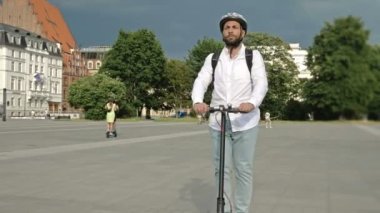 Sırt çantalı ve koruyucu kasklı sakallı bir adam şehir meydanında elektrikli scooter kullanıyor. Kullanışlı ve çevre dostu bireysel şehir taşımacılığı kavramı. 4K.