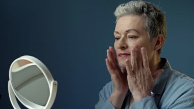Yaşlı, kır saçlı bir kadın aynadaki yansımasını dikkatlice inceler. Yüzündeki kırışıklıklara hafifçe dokunur. HD.