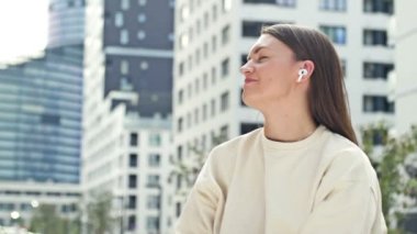 Kulağında kablosuz kulaklıkla müzik dinleyen çekici genç bir kadın. Modern Avrupa şehri, yaz günü. Kadın zevkten gözlerini kapar. HD.