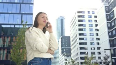 Modern bir Avrupa şehrinin caddesinde dururken akıllı bir telefonla konuşan neşeli genç bir kadın. HD.