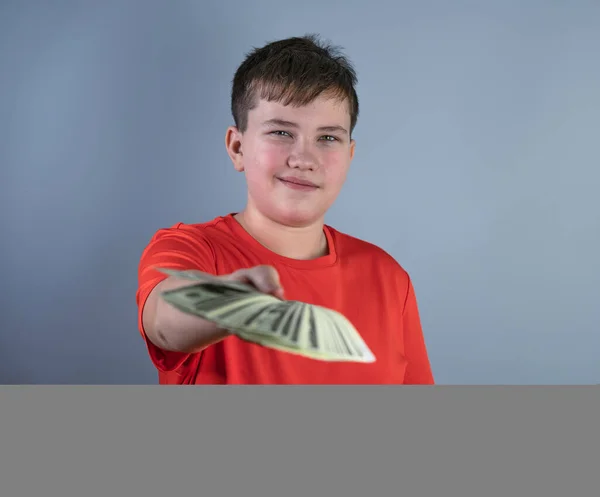 Ein Nahaufnahme-Foto eines zufriedenen Teenagers in einem roten T-Shirt, der Geldscheine in die Kamera hält. Ein junger Mann auf graublauem Hintergrund hält Dollars in der Hand — Stockfoto