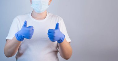 Beyaz tişörtlü ve maskeli genç bir adam elleri koruyucu mavi eldivenli bir baş parmak gösteriyor. Salgın sırasında yeni bir virüse karşı korunma kavramı, temiz eller. Boşluğu Kopyala