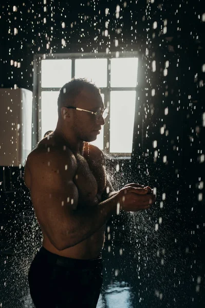 Handsome muscular man with glasses in shower. Young man with muscular torso taking shower, washing hair, standing under shower head with pouring water, caring for routine hygiene. Individuality.