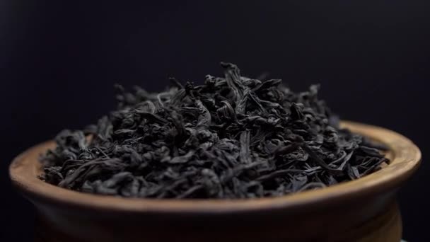 Close up widok suche czarne liście herbaty obracają się w glinianej misce na czarnym tle. Koncepcja - tradycyjna chińska herbata — Wideo stockowe