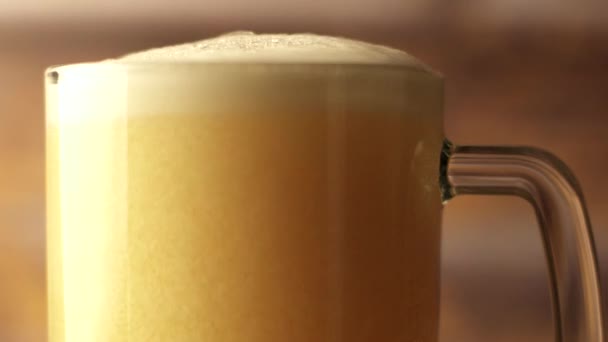 Close-up shot of beer is poured into a beer glass with a handle, a lot of bubbles and foam that flows down the glass. Warm wooden background — Stock Video