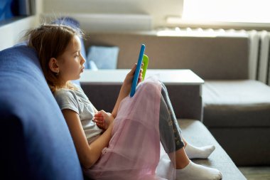 Kanepede oturan sevimli küçük kız teknolojiye bağımlı çocuk dijital tablet bilgisayarda online oyun oynamaktan zevk alıyor uygulamaları kullanıyor, evde internet sörfü bilgisini kullanıyor.,