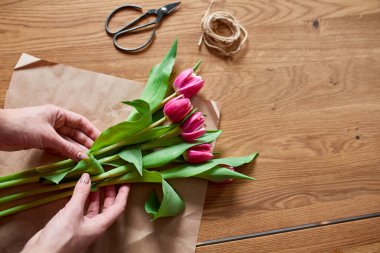 Ahşap masa üzerinde pembe lale buketi düzenleyen kadın elleri, çiçeklerle dolu iş yeri, iş yeri, diy, bahar hediyesi konsepti yukarıdan.