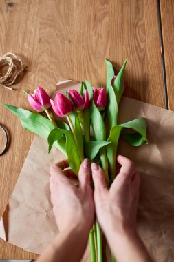 Ahşap masa üzerinde pembe lale buketi düzenleyen kadın elleri, çiçeklerle dolu iş yeri, iş yeri, diy, bahar hediyesi konsepti yukarıdan.