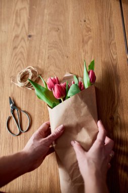 Ahşap masa üzerinde pembe lale buketi düzenleyen kadın elleri, çiçeklerle dolu iş yeri, iş yeri, diy, bahar hediyesi konsepti yukarıdan.