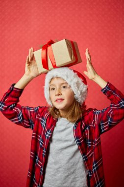 Noel Baba şapkalı şirin bir kız. Kafasında Noel hediyesi var. Kırmızı stüdyo arka planında.