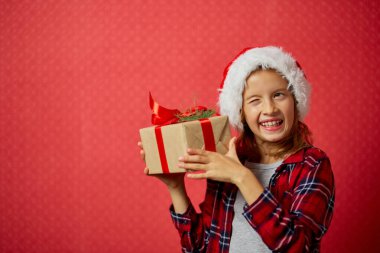 Noel Baba şapkalı, sürpriz ve pozitif küçük kız elinde Noel hediyesi ve hediye kutusunu işaretliyor, kırmızı arka planda izole edilmiş, mutlu yıllar.