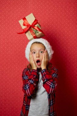 Noel Baba şapkalı şirin bir kız. Kafasında Noel hediyesi var. Kırmızı stüdyo arka planında.